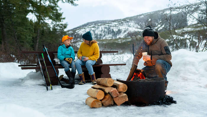 A family sitting outside in the winter with a firepit, stack of logs, and picnic table