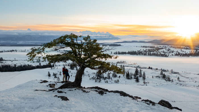 A mountain view of a couple standing by a tree at sunset with snow covering the ground.