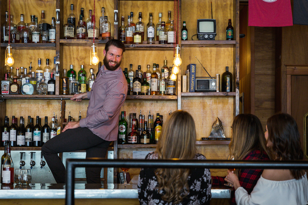 A bartender on a ladder in front of a shelf of liquor with three women sitting at the bar